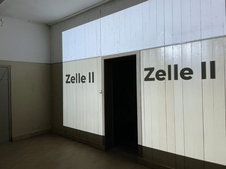 A dimly lit room with off-white walls, a doorway in the center, and the words Zelle II projected on both sides of the doorway.