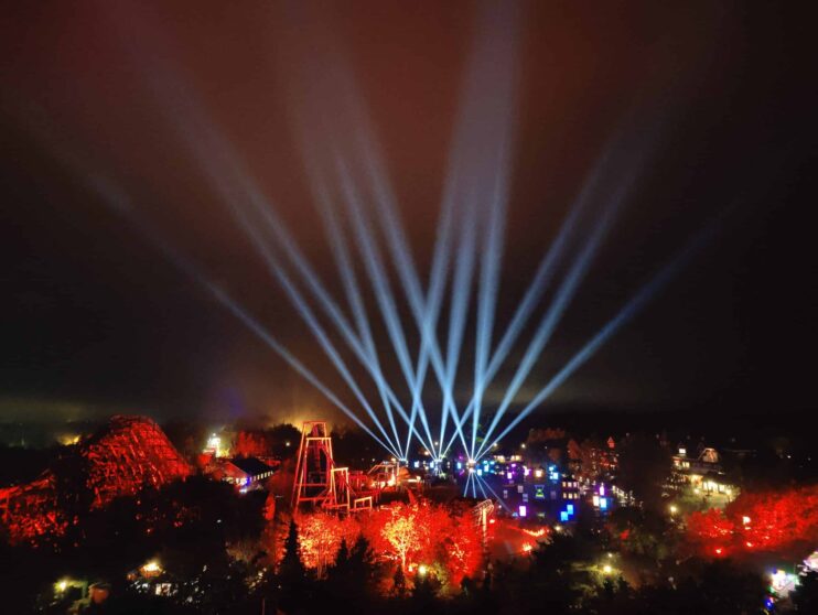 Bright white spotlights fan out into the night sky above a vibrant, colorfully lit outdoor festival with red and blue lights illuminating trees and structures.