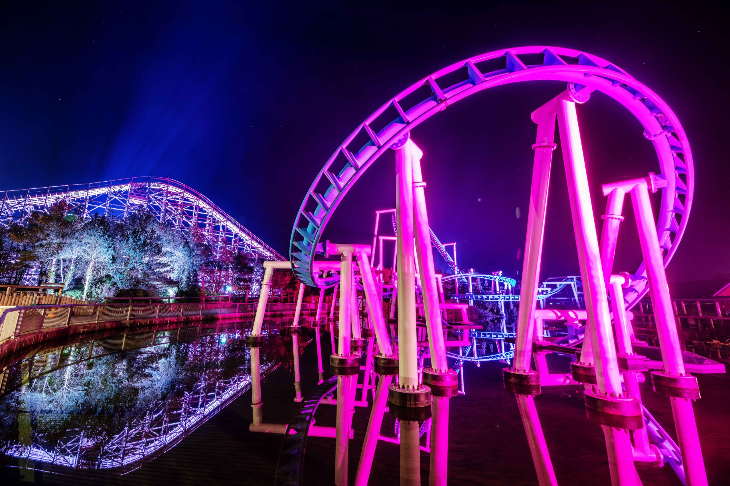 A brightly lit rollercoaster with purple and pink lights twists over water at night, with another coaster and trees in the background, reflecting vivid colors.