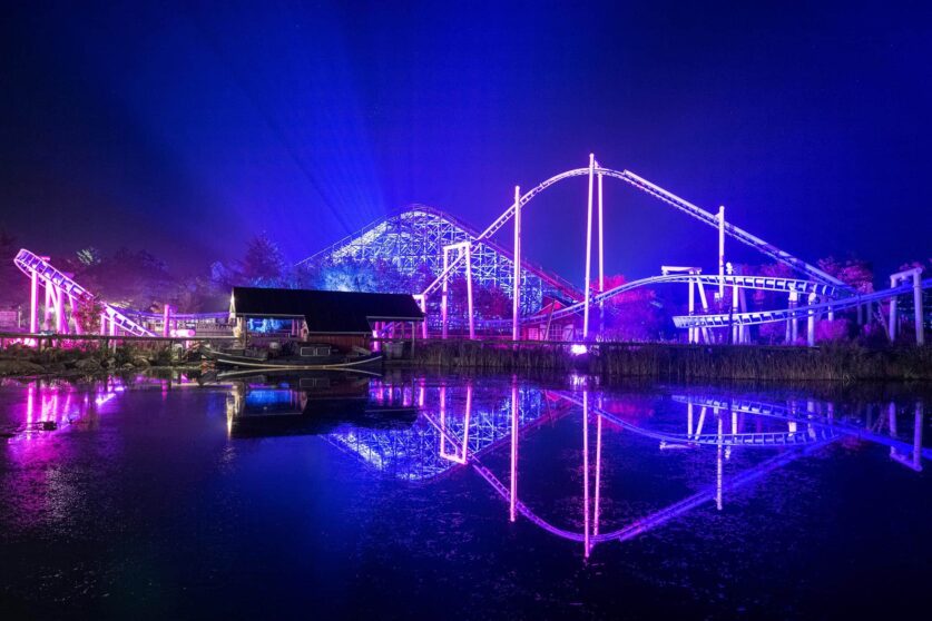 A brightly lit roller coaster at night glows with purple and blue lights, reflecting on a calm body of water in the foreground. A small wooden building stands near the ride, surrounded by trees.
