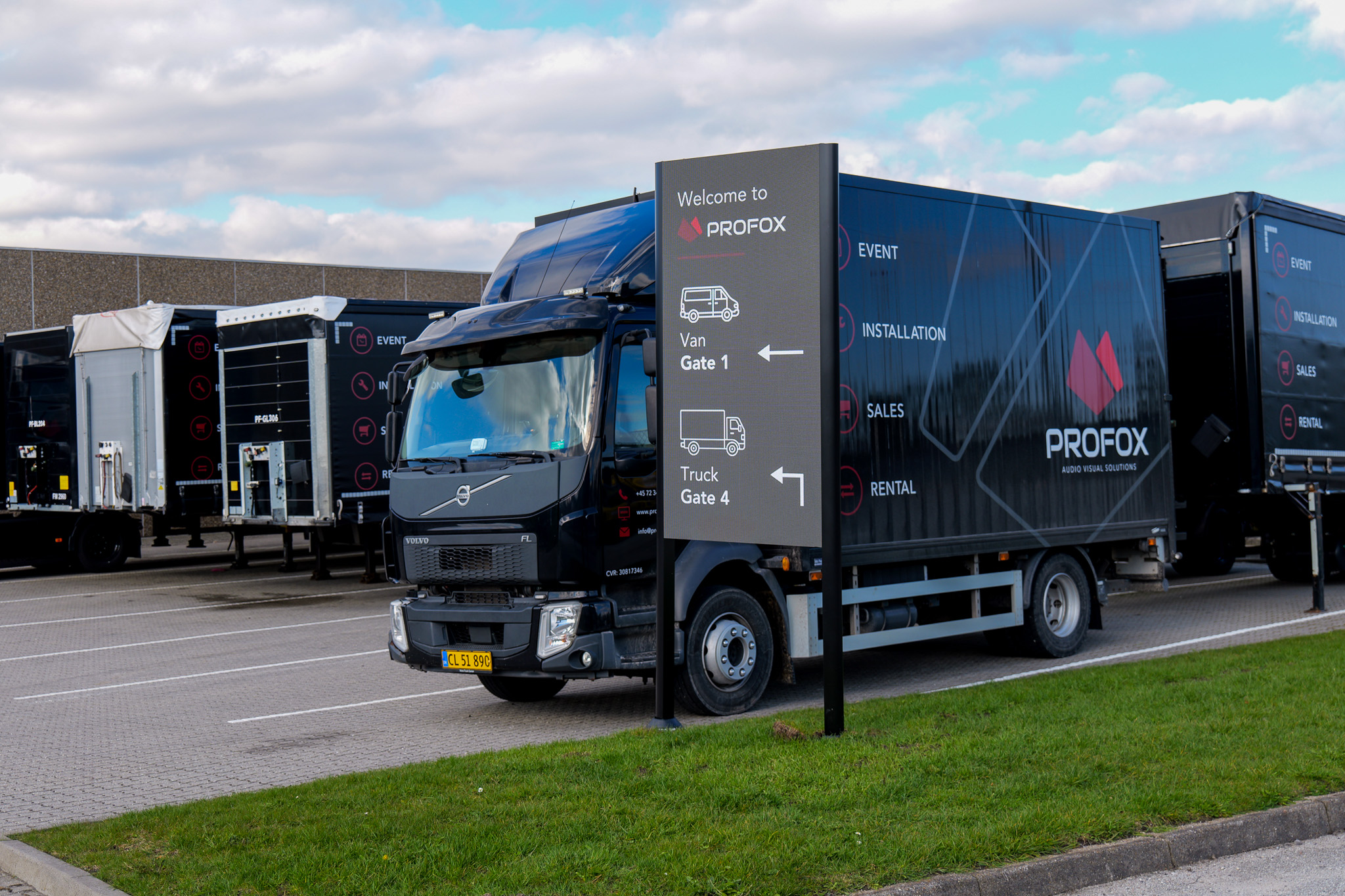 A PROFOX truck is parked near a sign showing directions for van and truck gates. Other trucks are parked in the background. The sky is partly cloudy.