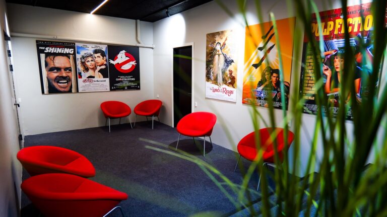 A small room with five red chairs, movie posters on white walls, gray carpet, and green plant leaves in the foreground. The posters include The Shining, Ghostbusters, and Pulp Fiction.