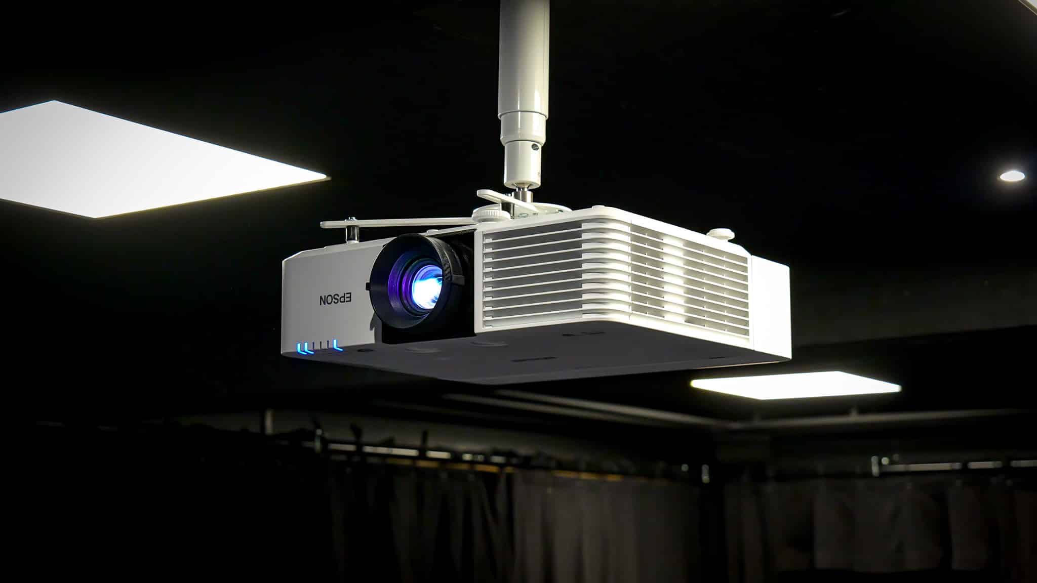 A ceiling-mounted white projector is turned on in a dark room, projecting blue light. Black curtains and illuminated ceiling panels are visible in the background.