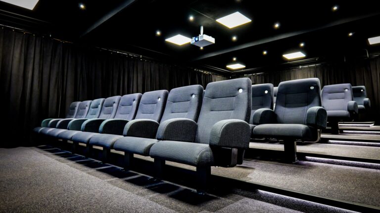 Rows of empty, modern, dark gray theater seats in a small cinema room with black walls, ceiling lights, and a ceiling-mounted projector.