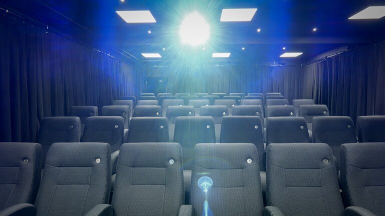 Rows of empty gray theater seats face a bright projector light in a dark, curtained cinema room.