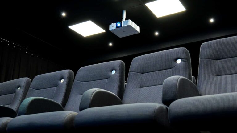Rows of empty, dark gray theater seats face a ceiling-mounted projector in a dimly lit room with square ceiling lights.