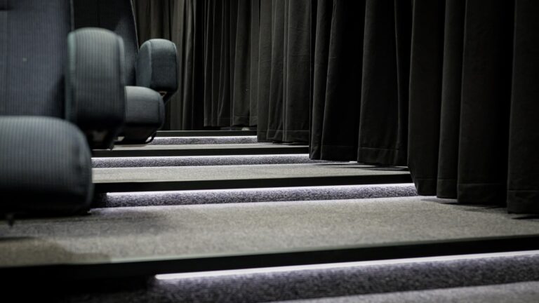 Rows of empty, dark fabric theater seats beside illuminated steps, with black curtains hanging alongside the aisle.