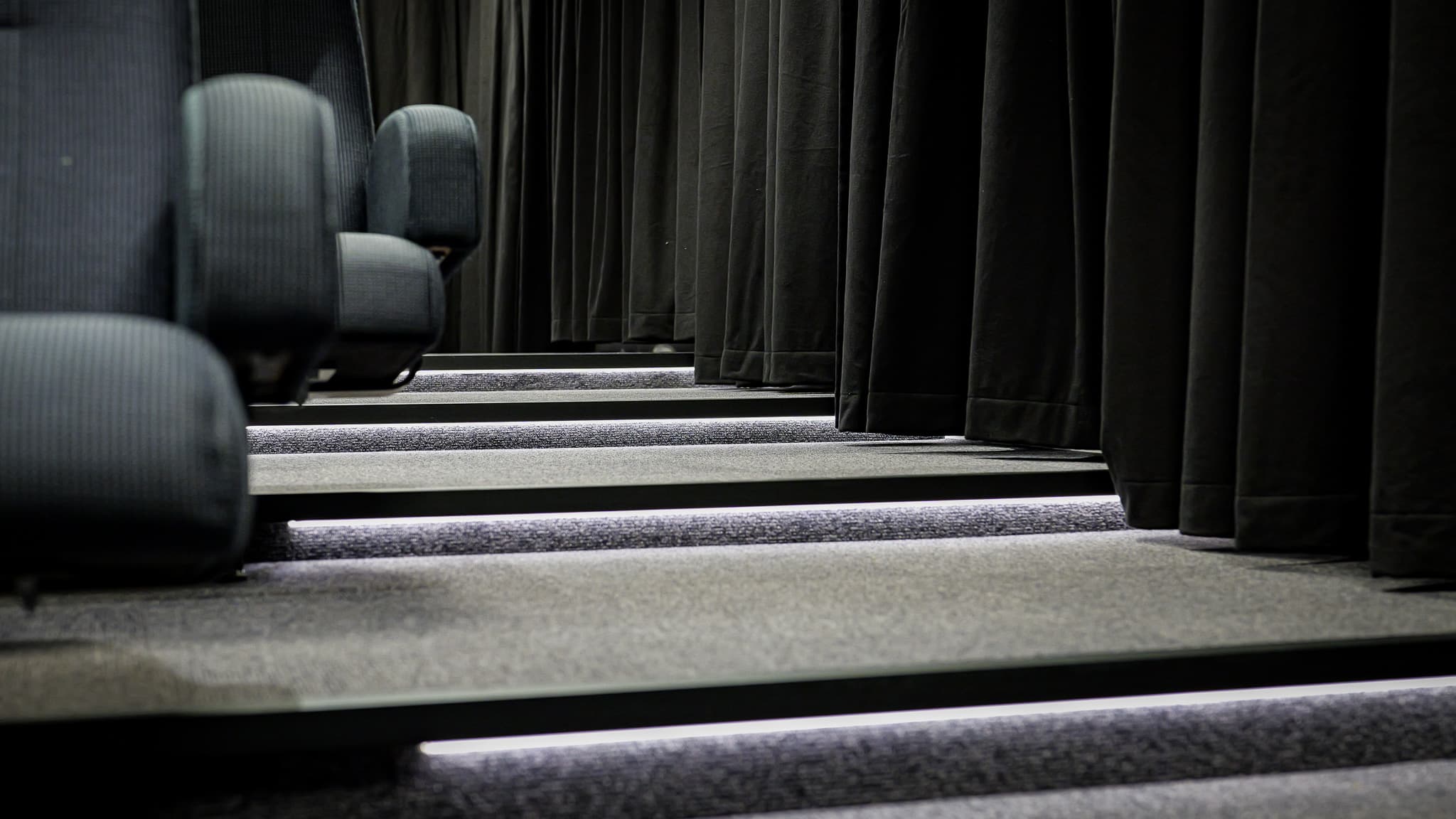 Rows of empty, dark fabric theater seats beside illuminated steps, with black curtains hanging alongside the aisle.