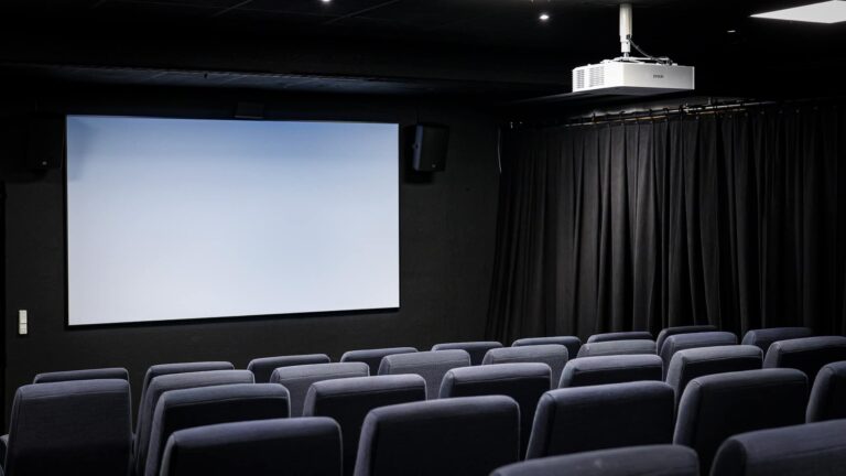 An empty movie theater with rows of gray seats faces a blank projection screen. Black curtains and a ceiling-mounted projector are visible, creating a modern, minimalist cinema atmosphere.