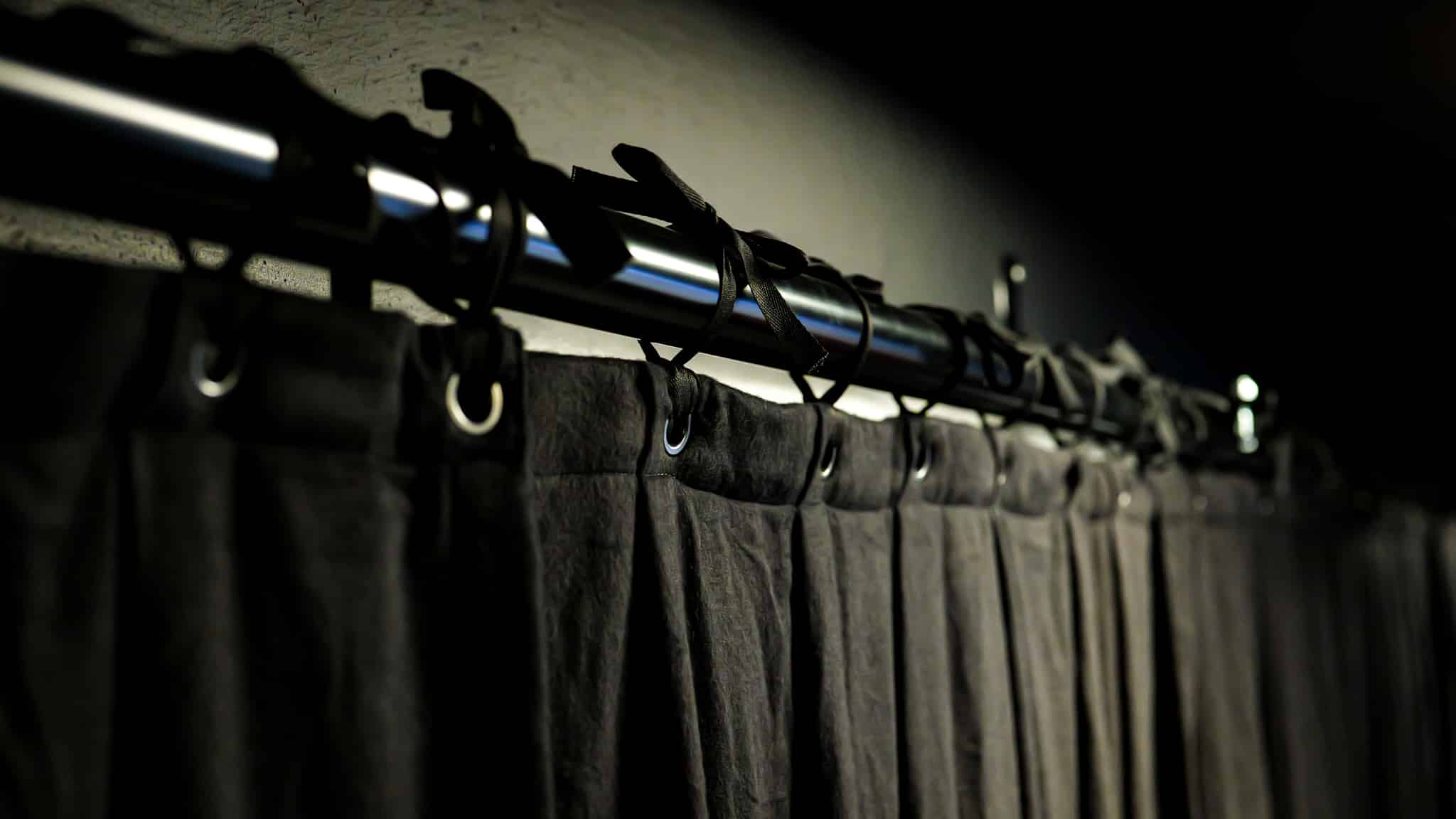 A close-up view of dark curtains hanging from metal rings on a shiny curtain rod, against a dimly lit wall.