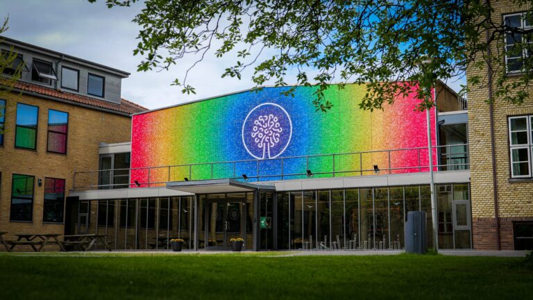 A modern building with a large, colorful rainbow mural featuring a white tree-like design above glass doors; trees and green grass frame the scene.