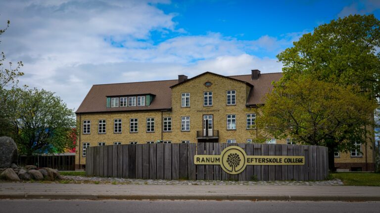 A large yellow-brick building stands behind a wooden fence with a sign reading Ranum Efterskole College. Green trees frame the scene under a partly cloudy blue sky.
