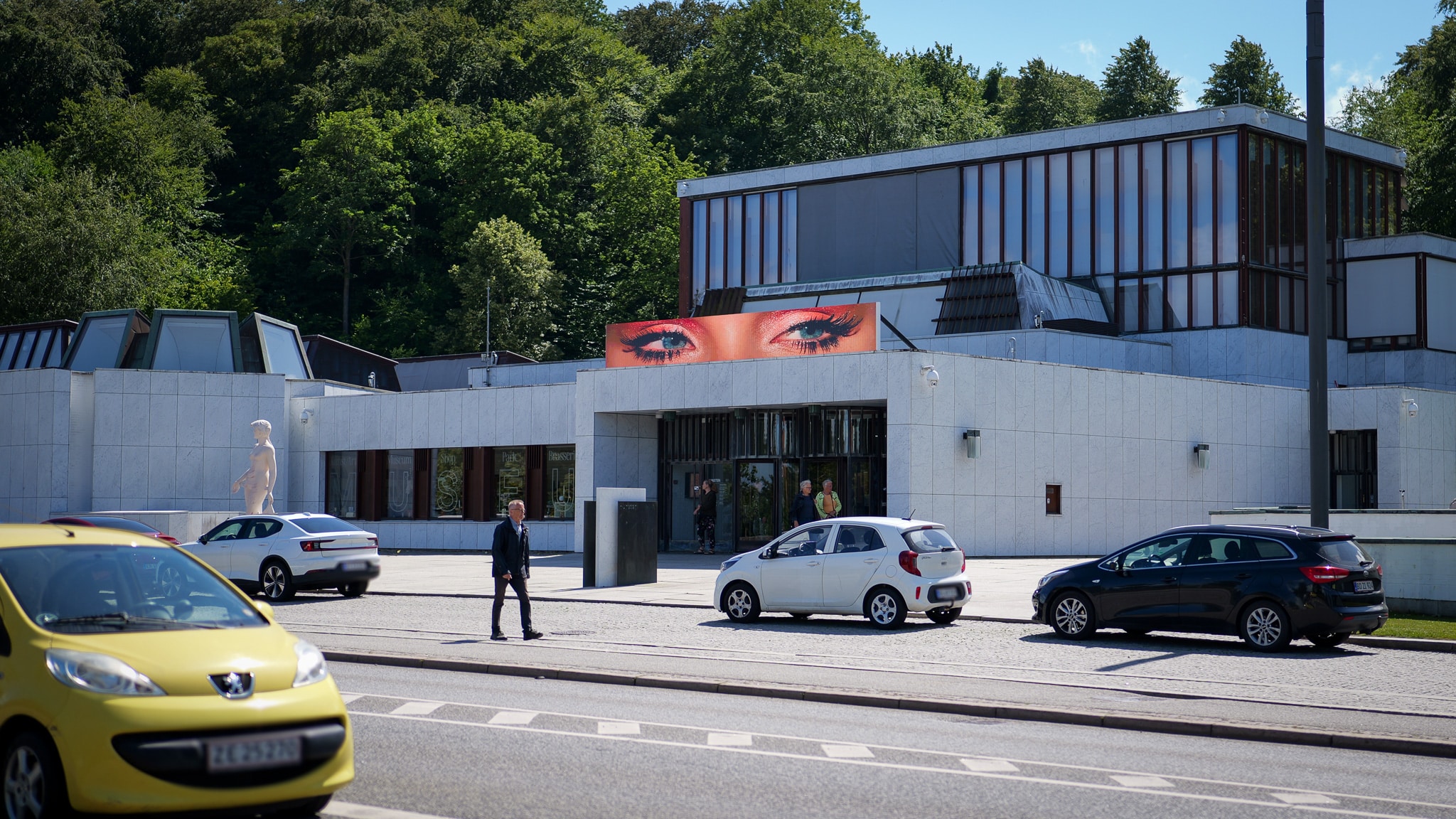 A modern white building with large windows features an art display of eyes above the entrance. Several cars are parked or driving by, and a man crosses the street in front. Trees fill the background.