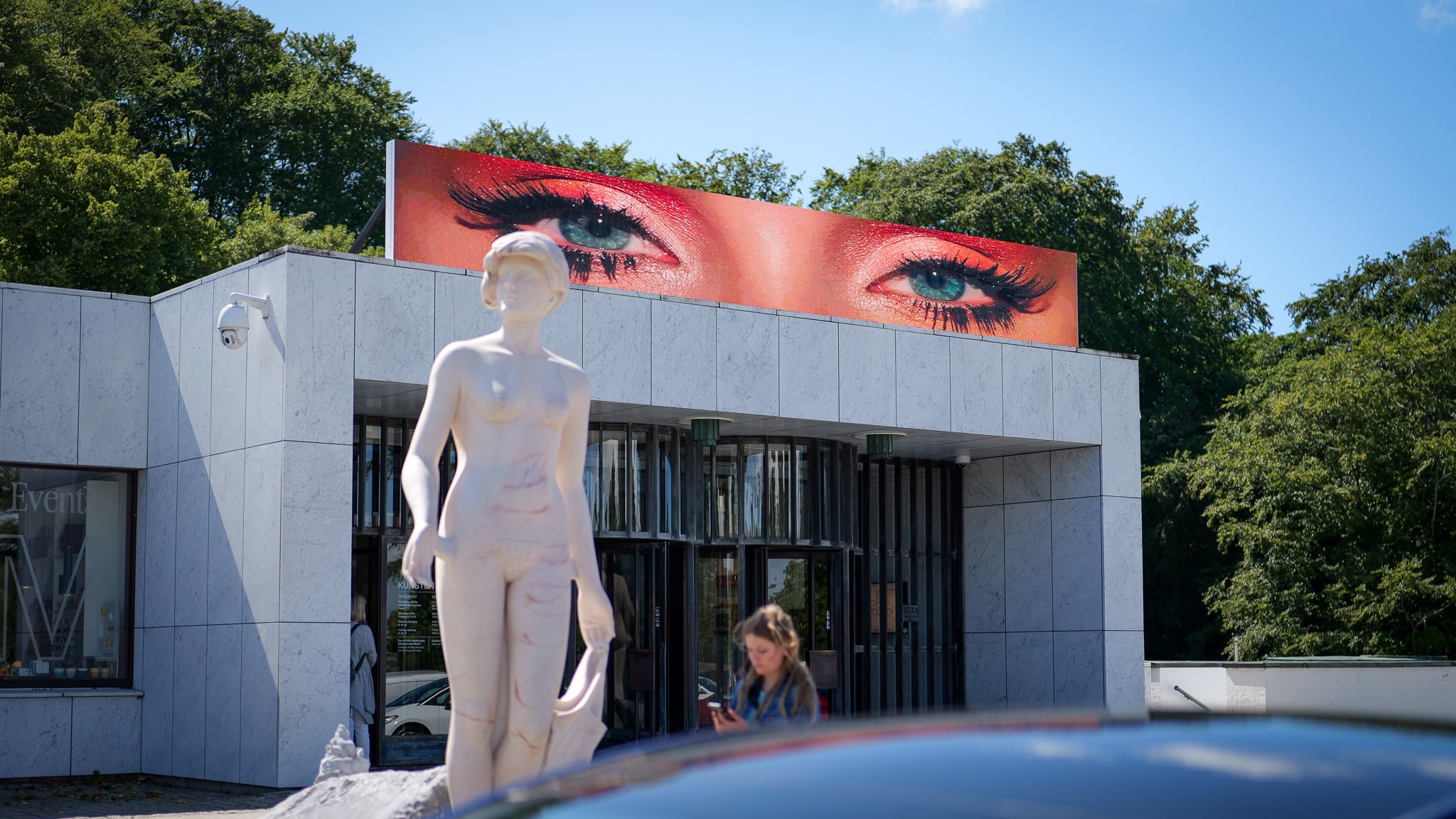 A white marble statue stands in front of a modern building with a large digital billboard displaying a close-up of made-up eyes above the entrance. Trees and blue sky are visible in the background.