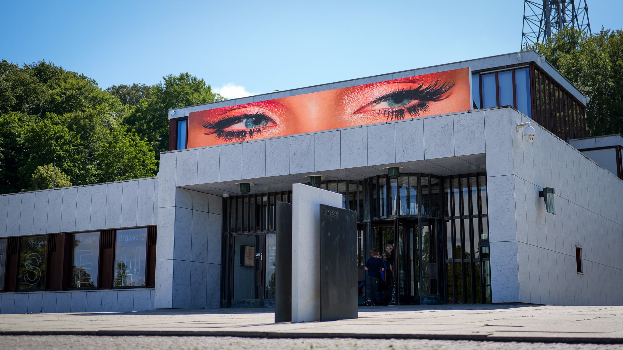 A modern building with large windows has a digital billboard on the roof displaying a close-up image of expressive eyes with bold makeup. Trees and blue sky are visible in the background.