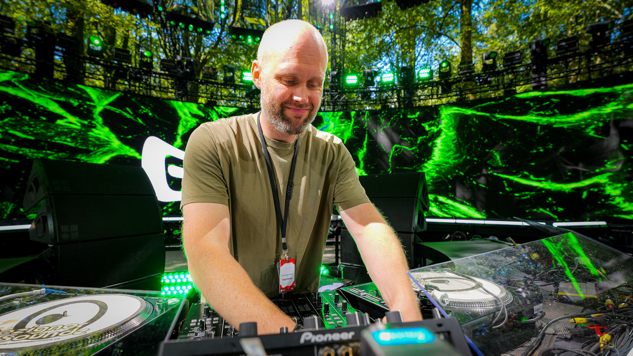 A DJ in a green t-shirt stands behind a Pioneer mixing console in front of a large EKKO digital screen.
