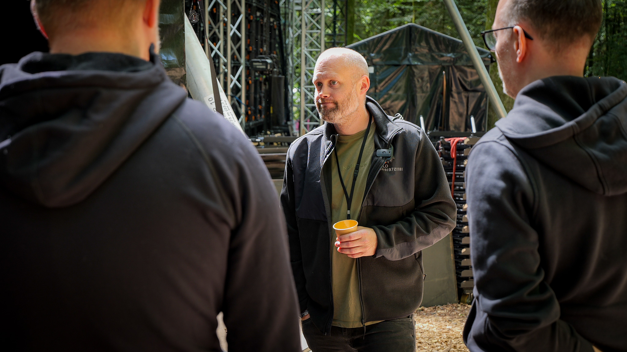 A man holding a yellow cup talks to two people with their backs to the camera at a Profox SFX outdoor event.