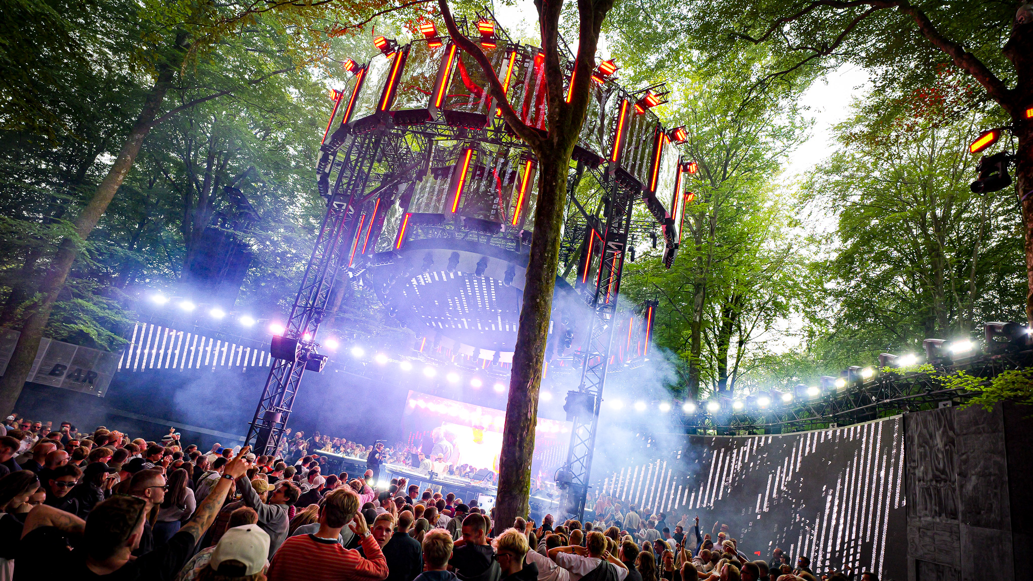 A large crowd faces a multi-tiered circular stage illuminated by LED panels, set against a backdrop of trees at an outdoor music festival.