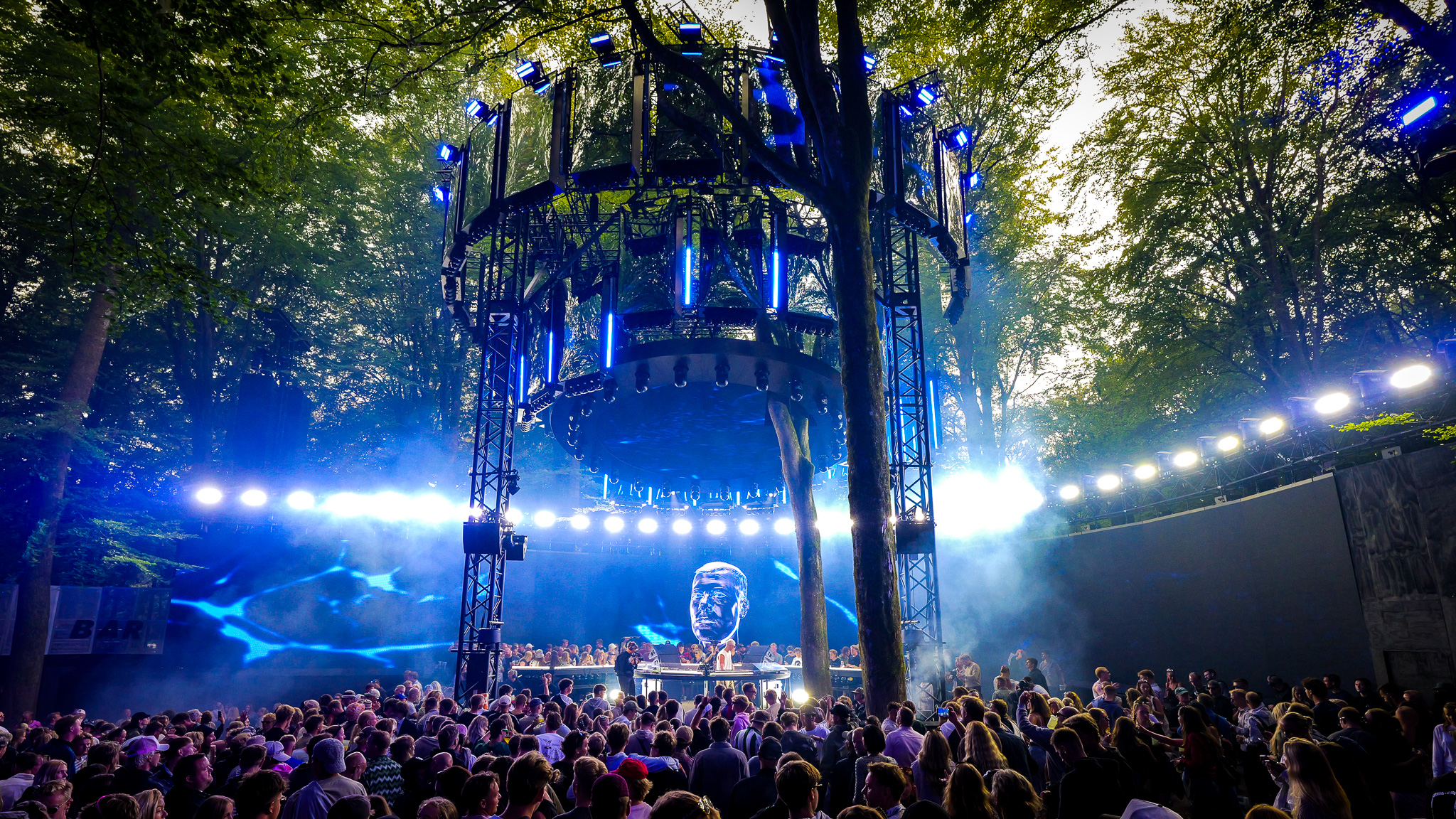 A large crowd gathers at an outdoor music festival featuring a towering, circular EKKO lighting rig suspended above a stage with a digital LED display of a face.