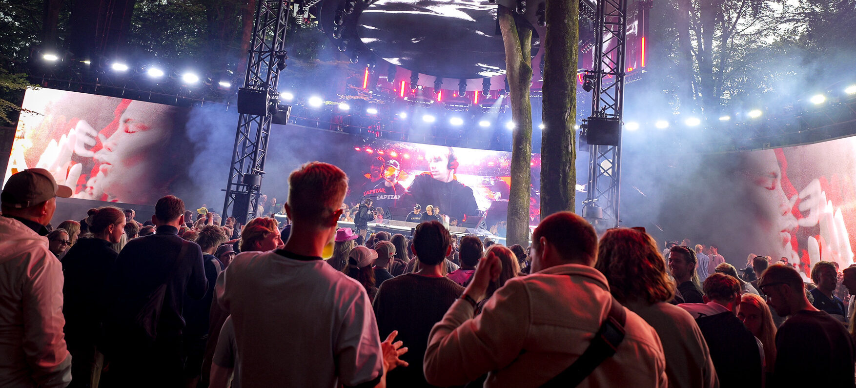 A crowd stands at an outdoor forest concert facing a large stage with Profox lighting and giant screens displaying a DJ performance with glowing red SFX.