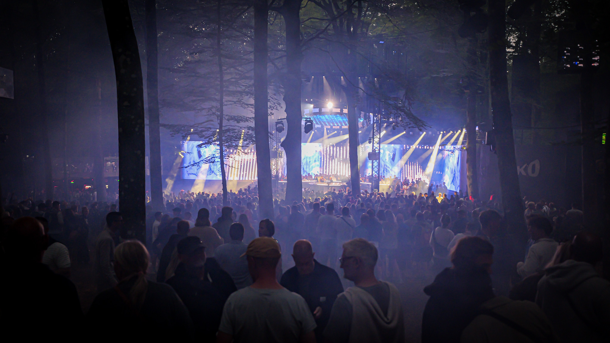 A large crowd stands in a forest at night, facing a music stage illuminated by bright light and a glowing video screen.