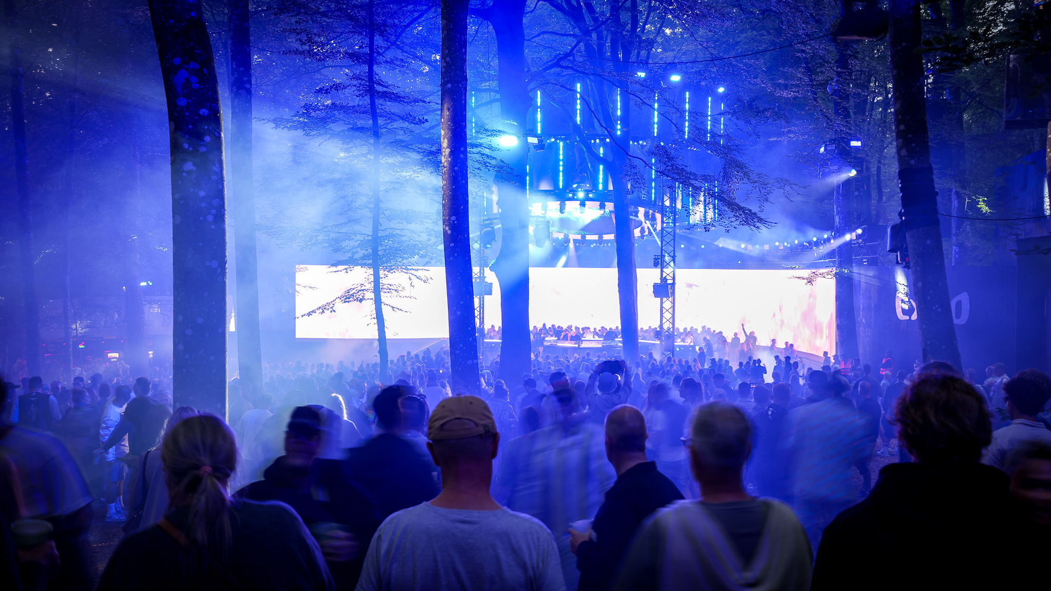 A large crowd watches a musical performance on an outdoor stage illuminated by blue LED lights and haze among forest trees.