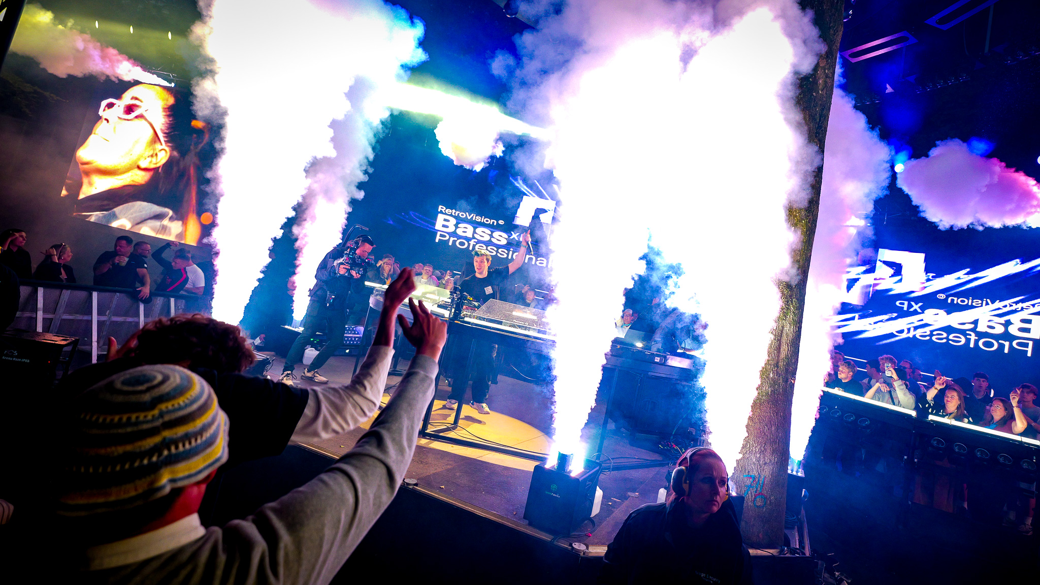 A DJ performs on an LED stage at a concert as two vertical columns of smoke rise in front of the light-filled crowd.