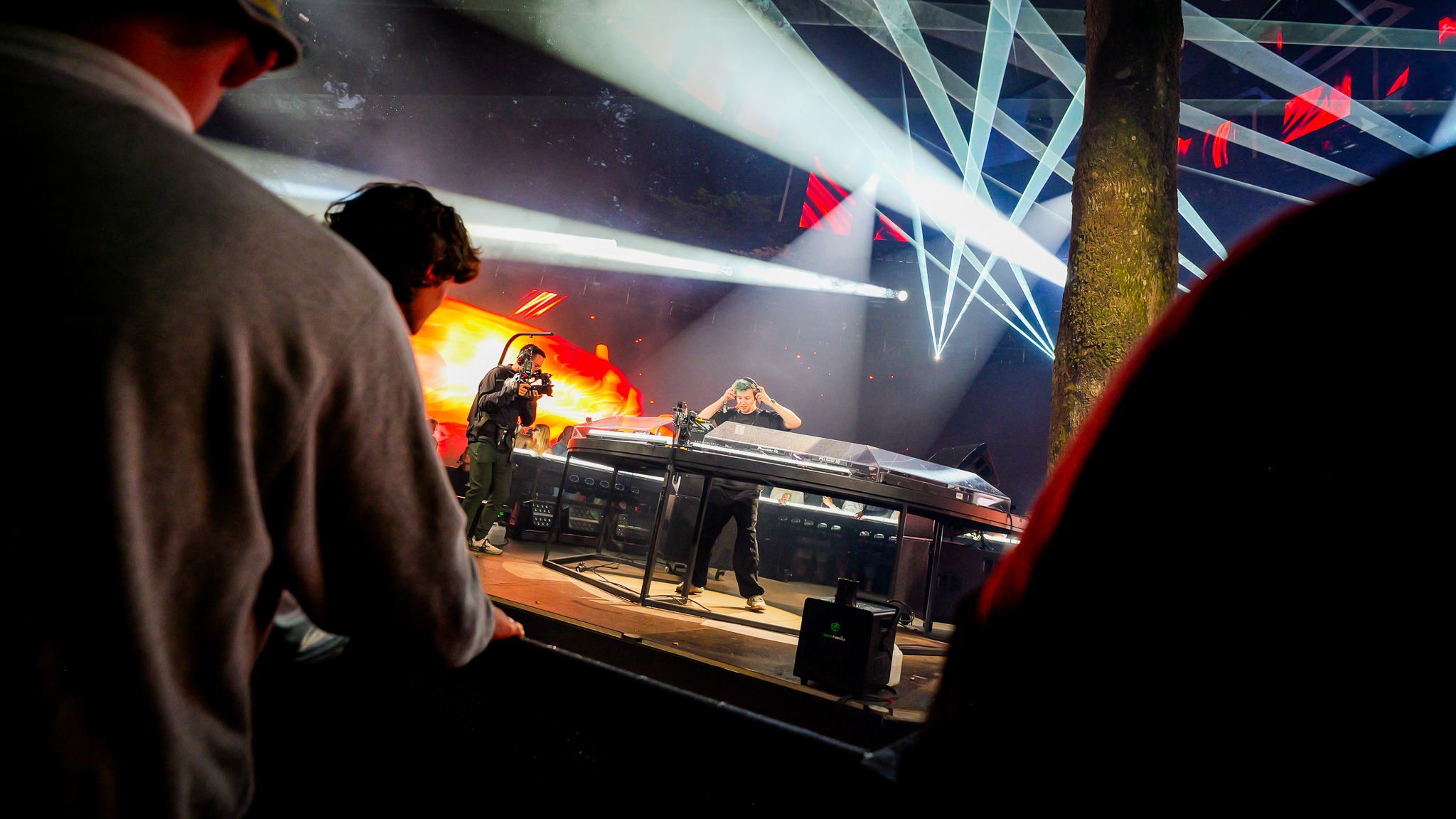 A DJ performs at a console under stage lights and a glowing LED screen, with a cameraman filming nearby and audience members in the foreground.