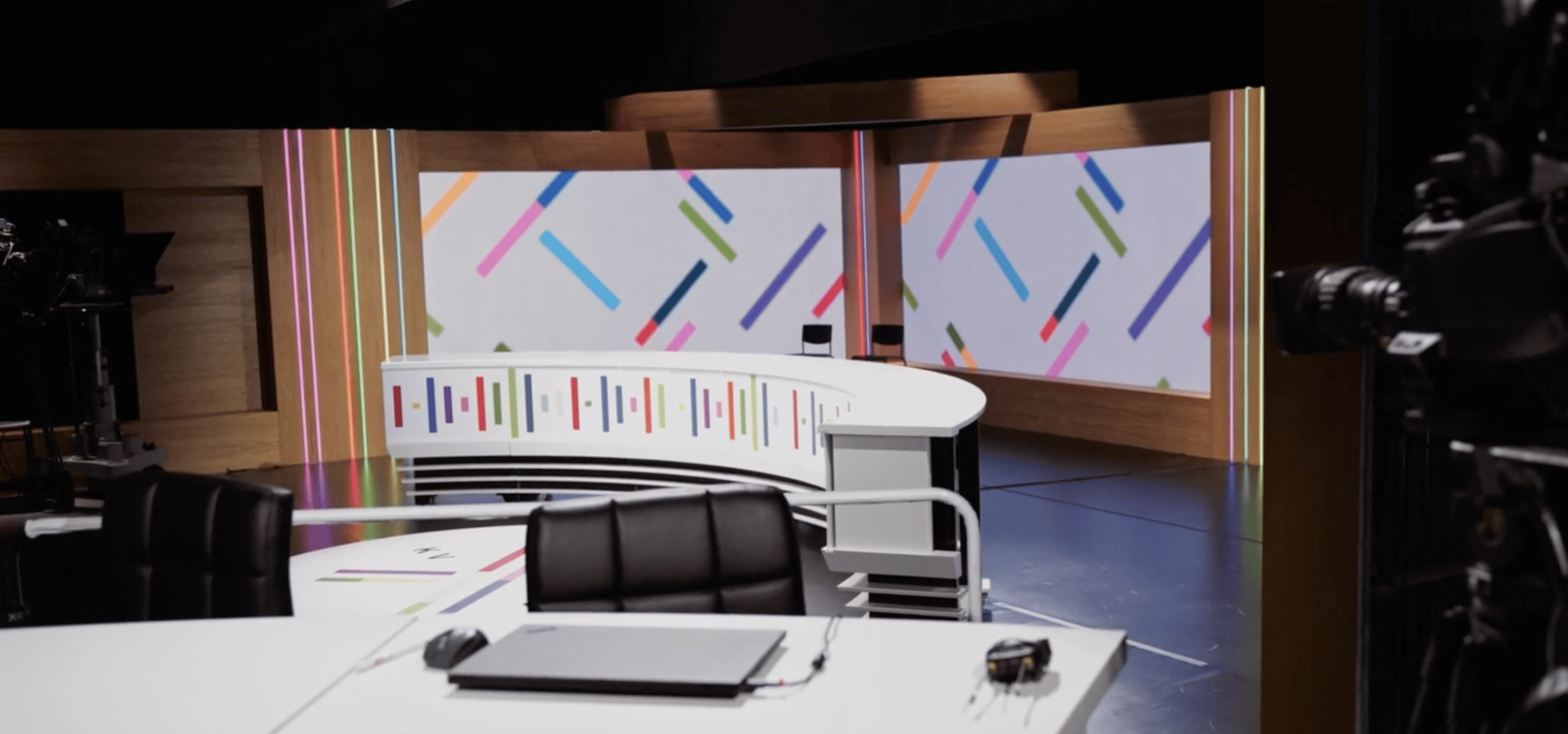 A modern TV studio set with curved white desks, black chairs, and colorful geometric patterns on wall panels in the background. Studio lights and cameras are partially visible.