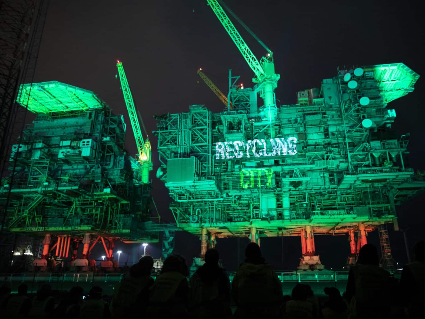 Two large offshore oil platforms illuminated in green lights at night, with the word RECYCLING glowing on one structure. Silhouetted people watch from the foreground.