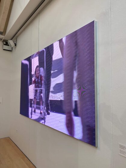 A video screen on a gallery wall displays a child in a stroller, with adults walking nearby. The image has a purple tint and is viewed from an angle.
