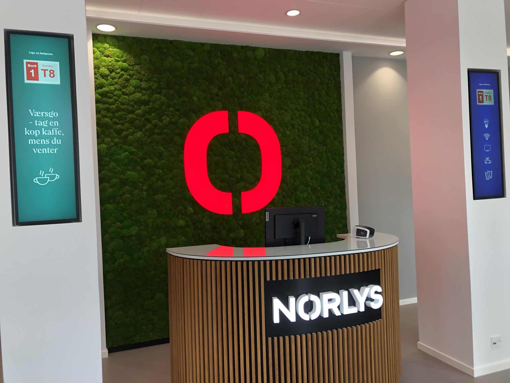 Reception desk with a NORLYS sign, a large red logo on a green moss wall behind, and two digital displays on either side showing information and coffee icons.