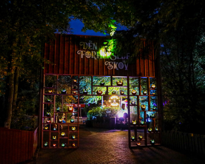 A wooden entrance decorated with glowing jack-o-lanterns at night. Colorful lights illuminate the trees and pathway beyond the gate, creating a festive Halloween atmosphere. The sign above reads DEN FORKJELSE SKOV..