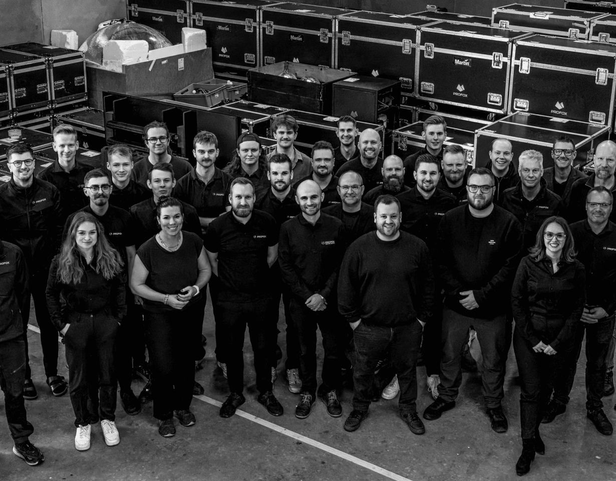 A large group of employees posing for a photo in a warehouse filled with stacked equipment cases labeled Profox and Martin.